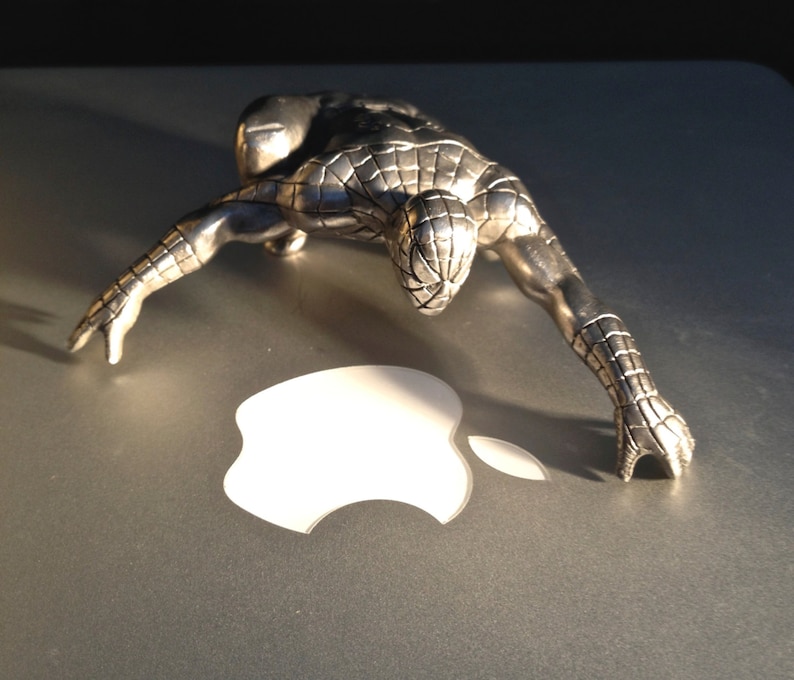 May include: A silver-colored Spider-Man figurine in a crouching pose. The figurine is detailed with textured lines and is positioned over a white Apple logo on a gray surface. The lighting creates shadows, highlighting the figure's form.