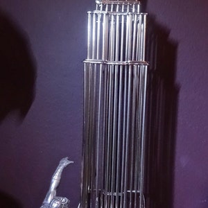 May include: A silver-colored metal sculpture of the Empire State Building with a Spider-Man figure climbing the side. The building is constructed of vertical bars, and the superhero is in a dynamic pose.