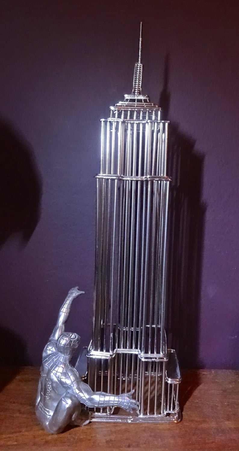 May include: A silver-colored metal sculpture of the Empire State Building with a Spider-Man figure climbing the side. The building is constructed of vertical bars, and the superhero is in a dynamic pose.