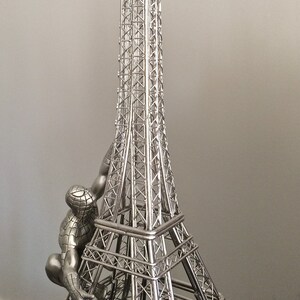 May include: A silver-colored Eiffel Tower figurine with a Spider-Man figure climbing the side. The tower is a detailed replica with intricate lattice work. The Spider-Man figure is in a crouching pose, adding a dynamic element to the sculpture.