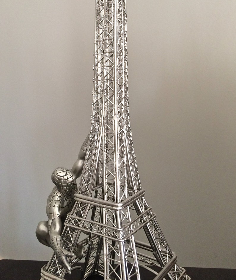 May include: A silver-colored Eiffel Tower figurine with a Spider-Man figure climbing the side. The tower is a detailed replica with intricate lattice work. The Spider-Man figure is in a crouching pose, adding a dynamic element to the sculpture.