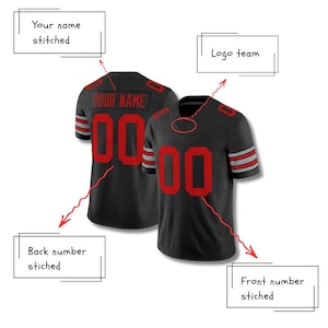 Puede incluir: Dos camisetas de f&uacute;tbol negras con n&uacute;meros y letras rojas. Las camisetas tienen rayas rojas y grises en las mangas. La parte trasera de una camiseta tiene el texto "YOUR NAME" cosido encima del n&uacute;mero 00. La parte delantera de la otra camiseta tambi&eacute;n tiene el n&uacute;mero 00.