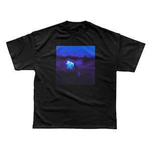 May include: Black t-shirt with a square graphic on the front. The graphic features a blurry, dark landscape under a blue sky, with a central, glowing white figure. The overall aesthetic is moody and atmospheric.