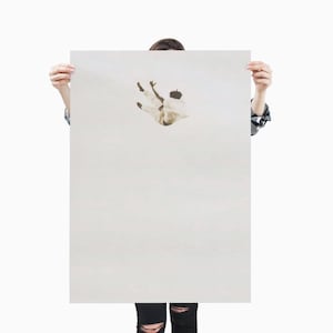 May include: A person holding a large, off-white poster with an artistic depiction of a figure in mid-air. The figure is wearing white clothing and appears to be falling. The background is a solid, light color.
