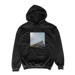 May include: Black hooded sweatshirt with a front pocket and a drawstring hood. The sweatshirt features a square graphic of a person standing on a building's edge against a blue sky. The hoodie is made of a soft, comfortable material.