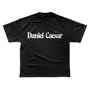 May include: Black t-shirt with the name "Daniel Caesar" printed in white, centered across the chest. The shirt has a crew neck and short sleeves. The fabric appears to be a soft, comfortable material.