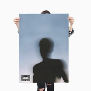 May include: A poster with a blurred silhouette of a person against a light blue background. The poster has a parental advisory warning in the lower left corner. The person is holding the poster.