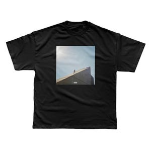 May include: Black t-shirt featuring a square image of a person standing on a building's roof against a blue sky. The image has a vintage aesthetic. The t-shirt is made of a soft material.