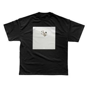 May include: Black t-shirt featuring a square graphic of a person falling. The graphic is in shades of white and gray, with the person's silhouette visible. The shirt is made of a soft material.