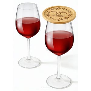 Puede incluir: Dos copas de vino transparentes llenas de vino tinto. Una copa tiene un posavasos de madera con el texto "Happy Birthday Juel" y un diseño de hojas. La otra copa está vacía.