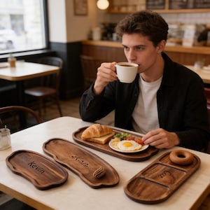Puede incluir: Bandejas de madera con nombres personalizados: Kevin, Kristie y Cace. Una bandeja contiene un desayuno de croissant, huevo frito y ensalada. Una persona está tomando café en una mesa en un café.