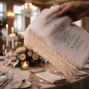 Può includere: Una pila di menu di nozze in lino bianco sporco con bordi sfilacciati viene tenuta in mano, rivelando un'elegante calligrafia. Il menu include i nomi "Alexandria", "Tomato Burrata Salad", "Alaskan Salmon" e "Prime Filet". Lo sfondo mostra una tavola ben apparecchiata.
