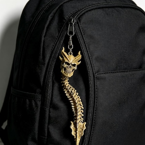 May include: A detailed, tan-colored skull and spine keychain hanging from a black backpack zipper. The keychain features a skull with horns and a spine that curves downward. The backpack is made of a black, textured material.