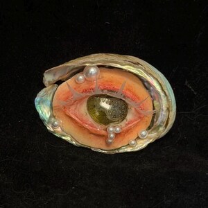 May include: A unique art piece featuring a sculpted eye within an iridescent seashell. The eye has a green iris, delicate lashes, and is surrounded by small pearls. The artwork is set against a black background.