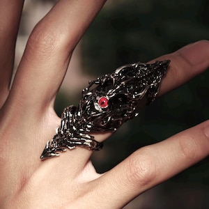Puede incluir: Anillo de metal oscuro con un diseño intrincado y esquelético. El anillo presenta una prominente gema roja, que se asemeja a un ojo, engastada en el detallado trabajo en metal. El anillo está diseñado para cubrir el dedo y el nudillo.
