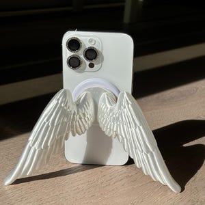 Könnte beinhalten: Weißes Smartphone mit einem dekorativen Handyhalter in Form von Engelsflügeln. Die weißen Flügel sind detailliert und erstrecken sich vom Telefon nach außen. Das Telefon hat drei Kameralinsen. Der Griff ist ein weißer Ring.