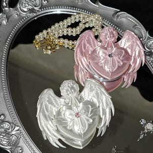 Könnte beinhalten: Zwei herzförmige Kompaktspiegel mit Engelsflügeln und Cherub-Figuren, einer in glänzendem Rosa und einer in perlmuttartigem Weiß, jeweils mit einem kleinen zentralen Edelstein. Eine Perlenkette mit goldfarbenen Anhängern und zwei kreuzförmige Ohrringe.