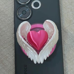 May include: A phone grip featuring a glossy, vibrant pink heart centered between two white, pearlescent angel wings. The accessory is attached to the back of a matte black smartphone, which displays a circular camera module at the top.