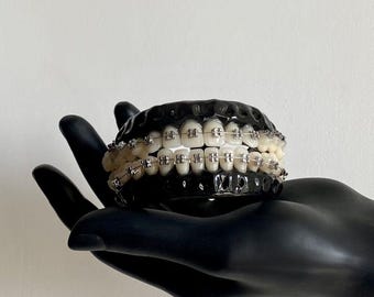 May include: A set of realistic human-like teeth featuring metallic orthodontic braces with silver brackets and wires, set within a glossy black gum-like base. The dental model displays a full upper and lower arch of white teeth with clear hardware.
