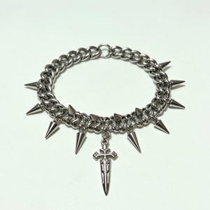 Könnte beinhalten: Ein silberfarbenes Choker-Halsband mit mehreren konischen Stacheln und einem Schwertanhänger. Die Halskette besteht aus dicken, ineinandergreifenden Kettengliedern. Die Stacheln sind gleichmäßig verteilt.