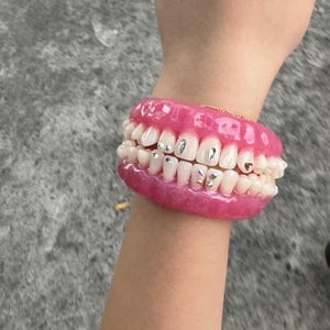 Puede incluir: Una pulsera circular que presenta un conjunto realista de dientes superiores e inferiores fijados en una base de resina rosa brillante. Varios dientes individuales están decorados con pequeños dijes metálicos plateados y cristales diminutos.