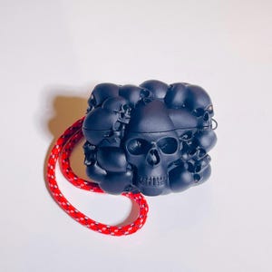 Può includere: Una custodia protettiva nero opaco per auricolari wireless caratterizzata da un motivo a rilievo tridimensionale di teschi multipli che coprono l'intera superficie. Un cordino in corda rosso e bianco maculato è fissato al lato.