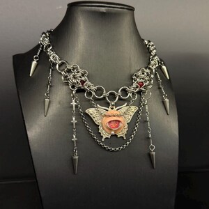 May include: A silver-toned necklace with a chainmail-style choker. The pendant features a butterfly design with a red eye-like center. Additional elements include dangling spikes and cross charms, displayed on a black mannequin.