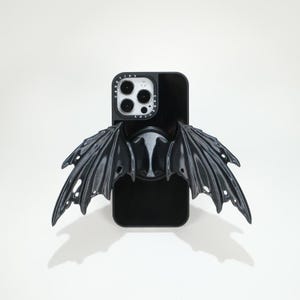 May include: Black phone case with attached black bat wings. The phone case has a glossy finish and a pop-up grip. The bat wings are detailed with a textured surface and a dark gray color.