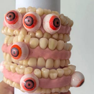 Könnte beinhalten: Eine zylindrische Skulptur mit mehreren Reihen realistischer, menschenähnlicher Zähne, die in ein zahnfleischfarbenes, rosa Material eingebettet sind, durchsetzt mit mehreren kugelförmigen Augäpfeln mit weißer Sklera, roten Iris und schwarzen Pupillen.