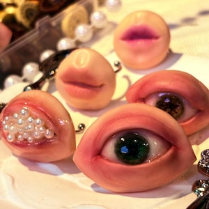 May include: A collection of sculpted anatomical art pieces including two realistic eyes with green irises, two sets of pale pink lips, and one eye-shaped sculpture filled with small white pearls, all featuring smooth, flesh-toned surfaces.