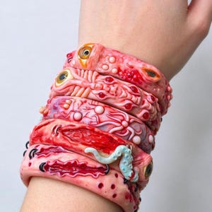 May include: A stack of five textured bracelets featuring a fleshy, organic aesthetic in shades of pink and red. The designs include embedded glass eyes, raised bumps, deep red crevices, black stitching details, and a pale blue tentacle accent.