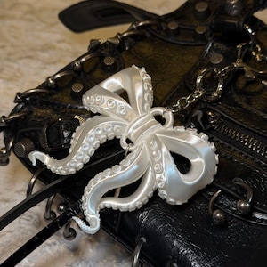 May include: A pearlescent white octopus charm with detailed tentacles hangs from a silver chain. The charm is set against a black leather bag with silver hardware, including rings and studs. The bag has a textured appearance.