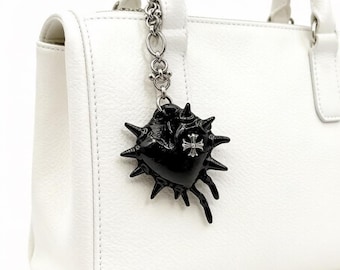 May include: A white leather handbag featuring a silver chain attachment with a black, spiked, heart-shaped charm. The charm is adorned with a small, metallic, cross-shaped emblem centered on its surface, contrasting against the white bag.