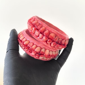 May include: Two circular bangles featuring a realistic, textured design of human teeth embedded into a vibrant red, gum-like base. The teeth are off-white with subtle shading, creating a detailed, anatomical appearance across the entire surface.