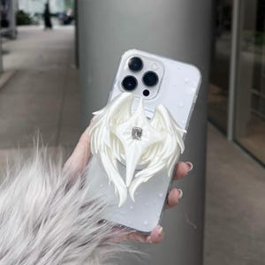 May include: A clear smartphone case featuring a white, three-dimensional wing-shaped phone grip attachment. The center of the wing design includes a small, rectangular, clear crystal accent. The case surface is decorated with subtle, small white star icons.