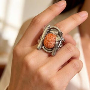 May include: A silver-toned ring featuring a miniature plate with a brain-shaped centerpiece, a knife, and a fork. The brain is a reddish-orange color. The ring is a unique piece of jewelry.