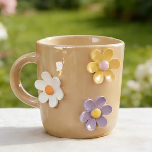 May include: A beige ceramic mug with a curved handle, adorned with three dimensional flower embellishments. The flowers are white with orange centers, yellow with pink centers, and purple with yellow centers. The mug is set against a blurred green background.
