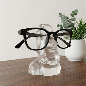 May include: A clear, transparent Moai statue eyeglasses holder with black-framed glasses resting on the nose. The statue is on a wooden surface, with a small potted plant in the background.