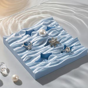 Puede incluir: Un expositor de joyas azul claro en forma de ola con varias joyas. El expositor contiene una perla, un broche en forma de copo de nieve y dos anillos. Los anillos tienen piedras transparentes. Un cristal transparente y una perla también están en la pantalla.