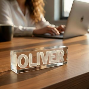 May include: A clear acrylic desk nameplate with the name "OLIVER" in white, set on a wooden desk. The name is in a bold, sans-serif font. A person is working on a laptop in the background.