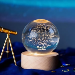 Op de afbeelding: Een heldere glazen bol met een sterrenbeeldkaart, van binnenuit verlicht, staat op een houten voet. De bol heeft het woord "Madrid" en de datum "2024-09-21" erop gedrukt. Een kleine messing telescoop staat links.
