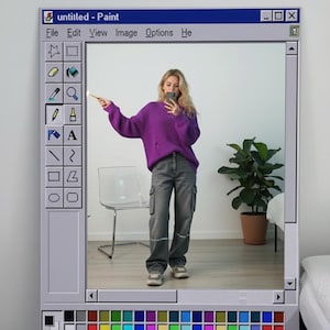 May include: A framed artwork styled as a Windows Paint interface, titled "untitled - Paint." The frame surrounds a person in a purple sweater and grey cargo trousers. A plant in a terracotta pot is visible in the background.