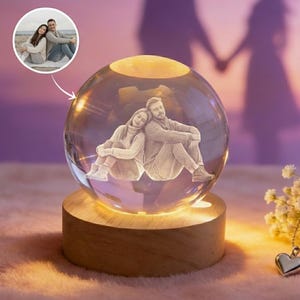 Op de afbeelding: Een heldere kristallen bol met een verlicht interieur, met een geëtste afbeelding van een stel. De bol staat op een ronde houten basis. Een kleine inset-afbeelding toont het paar. Het algemene thema is romantiek en relaties.