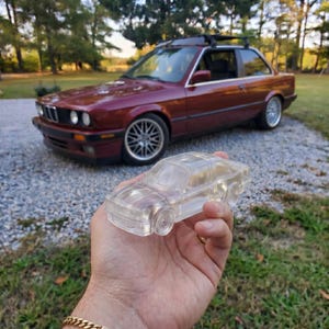 May include: A clear, transparent miniature car is held in front of a maroon-coloured vintage car. The miniature car is a detailed replica. The background features a gravel driveway and green grass.