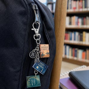 May include: A silver-toned metal keychain attached to a black backpack zipper, featuring a circular charm with the letter B and three miniature book charms titled The Mapmaker's Quest, Lights Over Harbor, and Songs of the Willow with detailed covers.