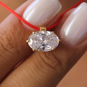 May include: A necklace featuring a clear, oval-cut gemstone set in a gold-toned metal prong setting. The pendant is suspended from a thin, vibrant red cord that encircles the neck, providing a striking contrast to the faceted, sparkling stone.