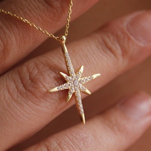 May include: A gold-colored necklace with a starburst pendant. The pendant is encrusted with small, clear gemstones. The necklace is displayed against a blurred background, highlighting the jewelry's details. The starburst design is a popular jewelry motif.