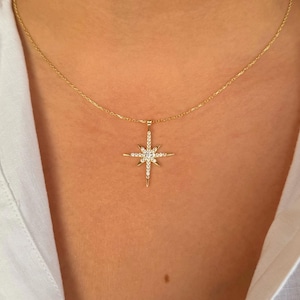 May include: A gold-colored necklace featuring a starburst pendant. The pendant is encrusted with small, clear gemstones. The necklace is worn against a white shirt.