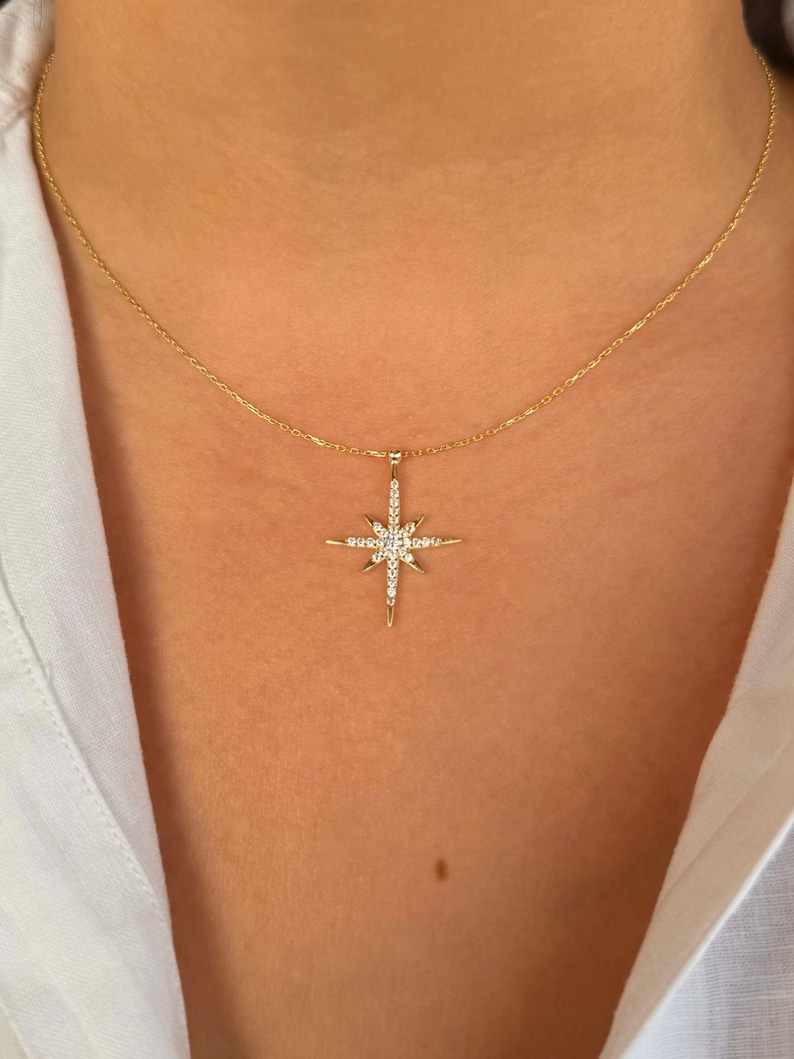 May include: A gold-colored necklace featuring a starburst pendant. The pendant is encrusted with small, clear gemstones. The necklace is worn against a white shirt.