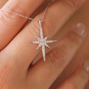 May include: A silver necklace featuring a starburst pendant encrusted with small, clear gemstones. The pendant is suspended from a delicate chain. The jewelry is displayed against a skin-toned background.
